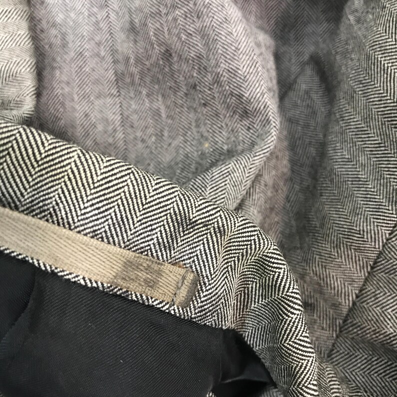 May include: Close-up of grey herringbone fabric with a black and white pattern. The fabric is folded, revealing a glimpse of a black lining and a beige waistband. The texture of the fabric is visible, showcasing the detailed weave.