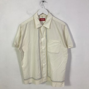 Può includere: Camicia color crema a maniche corte con logo Coca-Cola sul colletto. La camicia presenta strisce verticali blu e oro sul davanti e lungo l'orlo inferiore. Una singola tasca è sul petto sinistro.
