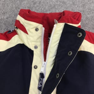 May include: Close-up of a vintage Fila jacket. The jacket features a color block design with red, cream, and navy blue panels. It has a white zipper and snap closures. The Fila logo is visible on the zipper pull.