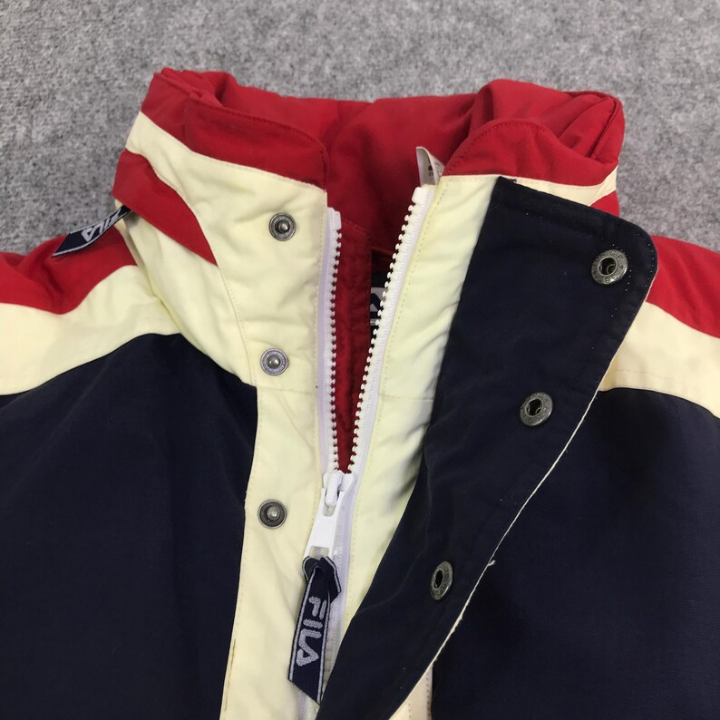 May include: Close-up of a vintage Fila jacket. The jacket features a color block design with red, cream, and navy blue panels. It has a white zipper and snap closures. The Fila logo is visible on the zipper pull.