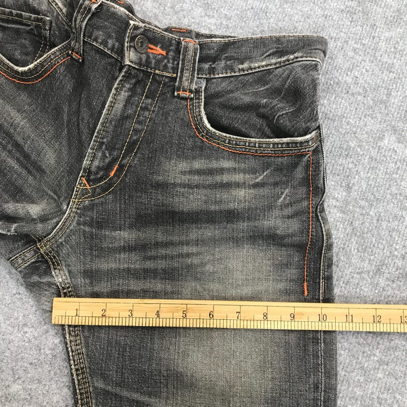May include: Close-up of a pair of dark gray denim jeans with orange stitching. The jeans have a button closure and pockets. A wooden ruler is placed across the jeans, measuring approximately 10 inches.