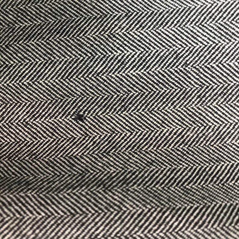 May include: Close-up of a black and white herringbone fabric. The fabric features a repeating chevron pattern, with alternating black and white stripes creating a textured appearance. The weave is tight, and the pattern is consistent across the surface.
