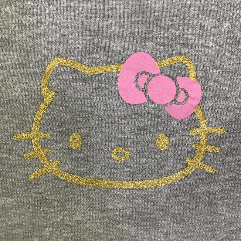 May include: A gray t-shirt featuring a gold glitter outline of a cartoon cat face with a pink bow. The cat's eyes and nose are also gold glitter. The design is centered on the shirt.