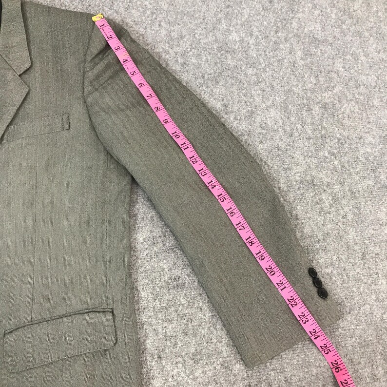 May include: A grey blazer with a notched lapel and a two-button closure. The sleeve is measured with a pink measuring tape, showing a length of approximately 66 cm. The blazer has a single pocket and is made of a textured fabric.
