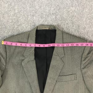 May include: A grey blazer with a black lining is measured with a pink tape measure. The blazer is approximately 46 cm wide. It features a notched lapel and two front pockets. The fabric has a subtle textured pattern.