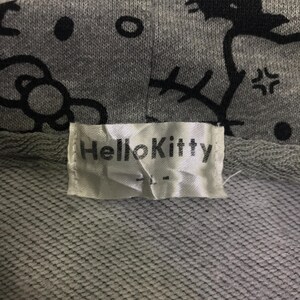 May include: Close-up of a grey hoodie featuring a black Hello Kitty pattern. The hoodie has a white label with the text "Hello Kitty". The fabric is a textured knit.