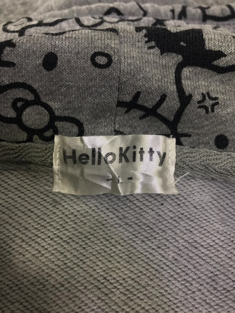May include: Close-up of a grey hoodie featuring a black Hello Kitty pattern. The hoodie has a white label with the text "Hello Kitty". The fabric is a textured knit.