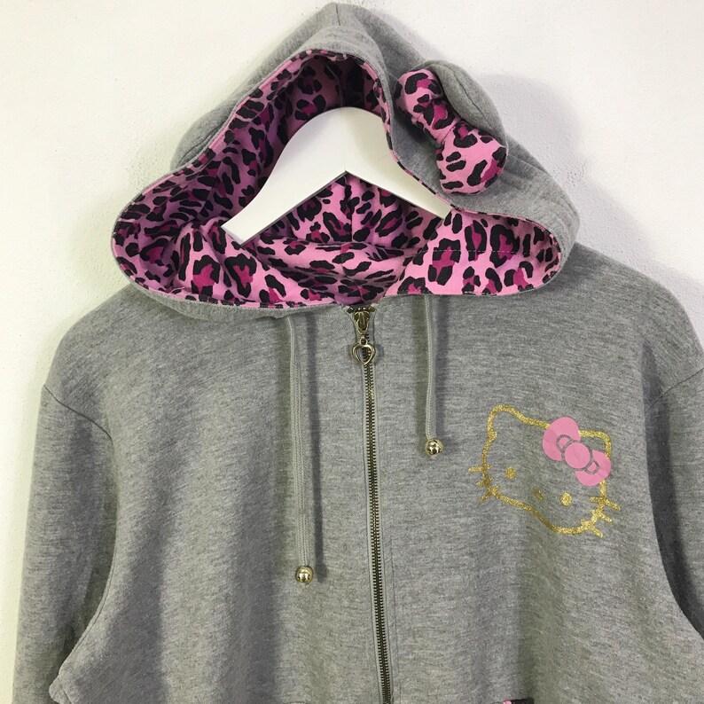 May include: A gray zip-up hoodie featuring a pink and black leopard print lining in the hood and ears. The hoodie has a gold glitter Hello Kitty graphic on the front.