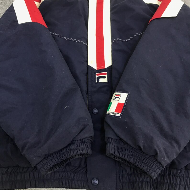 May include: Navy blue jacket with white and red accents. The jacket features a white stripe down the center with red stripes on the shoulders. The Fila logo is visible on the chest and sleeve. The jacket has a snap closure and elastic cuffs.