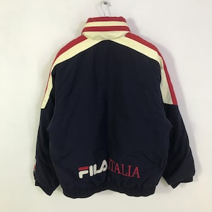 May include: A navy blue jacket with red and cream-colored accents. The jacket features the brand name "FILA" in white and the word "ITALIA" in red. The jacket has a high collar and is hanging from a metal hook.