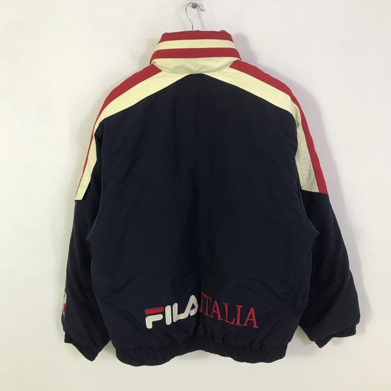 May include: A navy blue jacket with red and cream-colored accents. The jacket features the brand name "FILA" in white and the word "ITALIA" in red. The jacket has a high collar and is hanging from a metal hook.