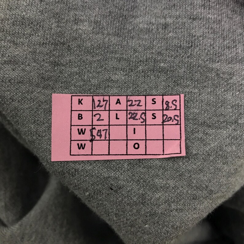 May include: A close-up of a gray fabric with a pink tag attached. The tag has handwritten numbers and letters in black ink. The fabric appears to be a soft material, possibly a sweatshirt or t-shirt.