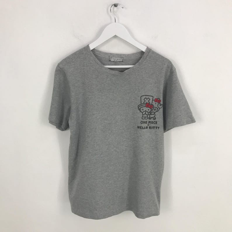 Pu&ograve; includere: T-shirt grigio a maniche corte con scollo a V. La maglietta presenta una piccola grafica sul davanti con la scritta "ONE PIECE HELLO KITTY". La grafica &egrave; un personaggio dei cartoni animati con cappello e fiocco.