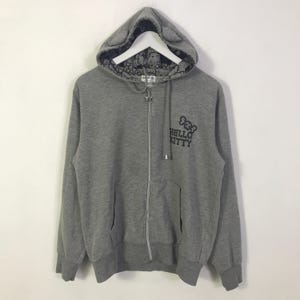 May include: A grey zip-up hoodie with a Hello Kitty design. The hoodie has a hood with cat ears and a patterned lining. The Hello Kitty logo is printed on the front. It features a front pocket and a star-shaped zip pull.