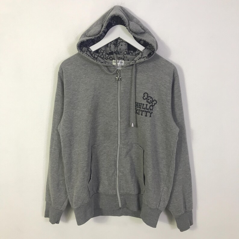 May include: A grey zip-up hoodie with a Hello Kitty design. The hoodie has a hood with cat ears and a patterned lining. The Hello Kitty logo is printed on the front. It features a front pocket and a star-shaped zip pull.