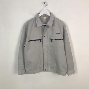 以下が含まれることがあります： ジッパー留めと2つのポケットが付いたライトグレーのワークジャケット。ジャケットの右胸には日立のロゴ、ポケットには赤、白、青のジッパーのディテールが施されています。