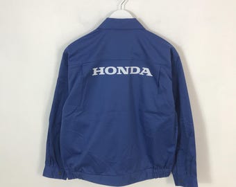 May include: A blue jacket with the word "HONDA" printed in white across the back. The jacket has a button-up front, long sleeves, and a collar. The jacket is hanging on a white hanger.