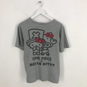 Pu&ograve; includere: Una maglietta grigia con una grafica di due personaggi dei cartoni animati con fiocchi rossi. Il testo "ONE PIECE x HELLO KITTY" &egrave; stampato sotto i personaggi in nero. La maglietta &egrave; appesa a una gruccia bianca.