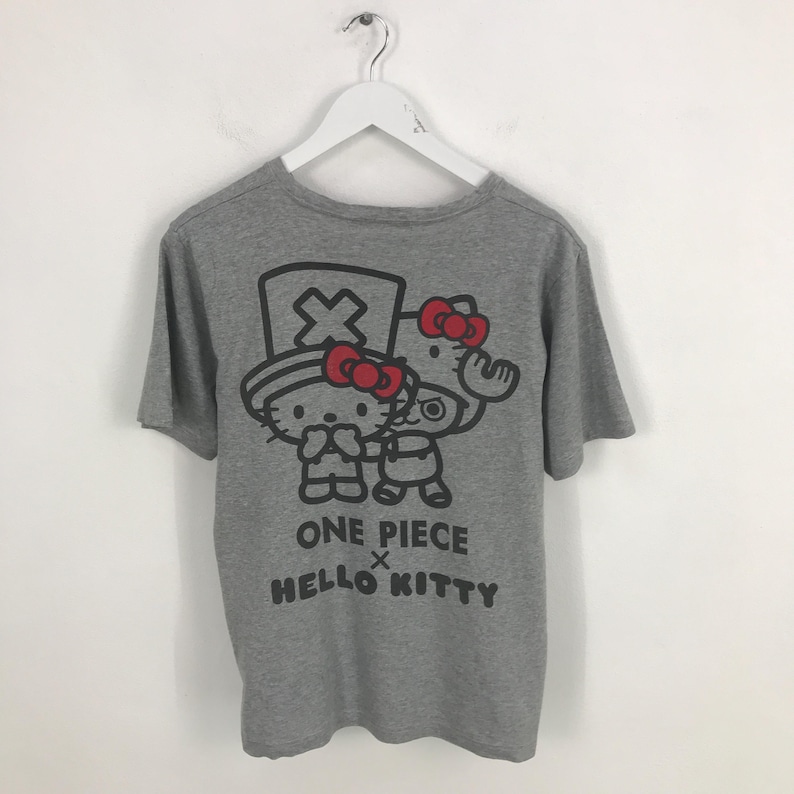 Pu&ograve; includere: Una maglietta grigia con una grafica di due personaggi dei cartoni animati con fiocchi rossi. Il testo "ONE PIECE x HELLO KITTY" &egrave; stampato sotto i personaggi in nero. La maglietta &egrave; appesa a una gruccia bianca.