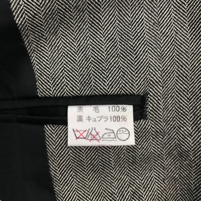 May include: Close-up of a black and white herringbone pattern fabric. A white tag with Japanese text and care symbols is visible. The fabric appears to be a wool blend.