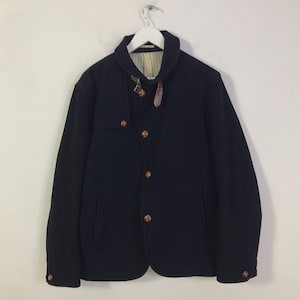 以下が含まれることがあります： ネイビーブルーのジャケット。ノッチドラペルカラーと木製ボタンが付いています。襟の近くに革ストラップのディテールがあるユニークなデザインです。白いハンガーにかかっています。