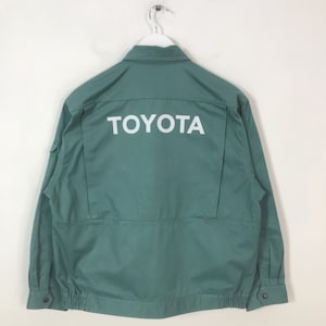 May include: Green work jacket with the word "TOYOTA" printed in white across the back. The jacket has a button-down front, a small pocket on the left sleeve, and elastic cuffs. The jacket is hanging on a white hanger.