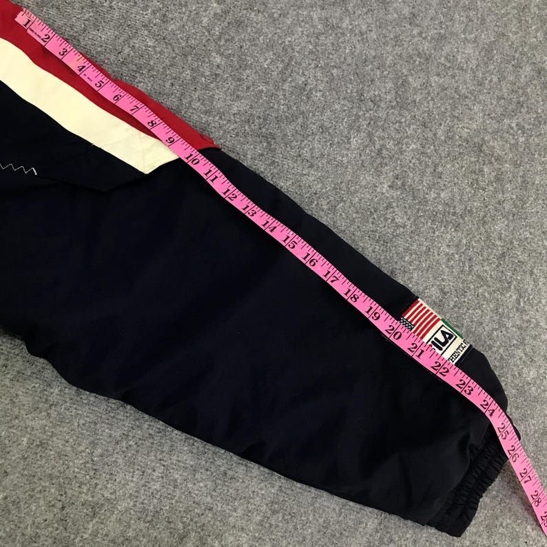 May include: A navy blue jacket sleeve with red and white accents. A pink measuring tape shows the sleeve is approximately 24 inches long. The jacket has a small patch with the brand name and a flag design.
