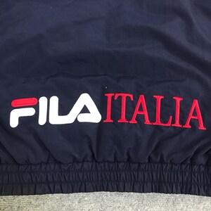 May include: Navy blue athletic jacket with the text "FILA ITALIA" in white and red. The jacket has a simple design and appears to be made of a smooth, possibly nylon, material. The text is embroidered on the jacket.