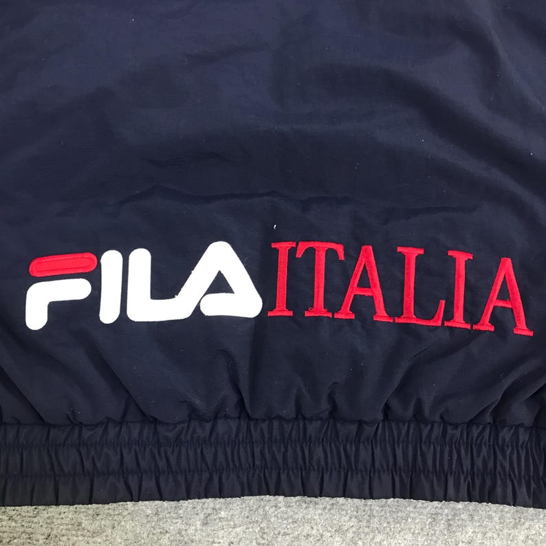 May include: Navy blue athletic jacket with the text "FILA ITALIA" in white and red. The jacket has a simple design and appears to be made of a smooth, possibly nylon, material. The text is embroidered on the jacket.