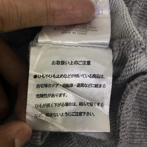 May include: A close-up of a grey fabric label with Japanese text and English text that reads "MADE IN CHINA". The label is attached to a grey sweatshirt. The text on the label provides care instructions.