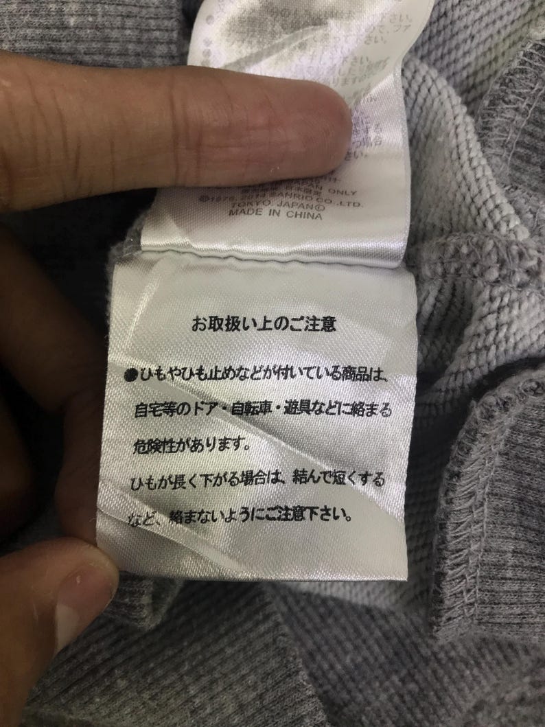 May include: A close-up of a grey fabric label with Japanese text and English text that reads "MADE IN CHINA". The label is attached to a grey sweatshirt. The text on the label provides care instructions.