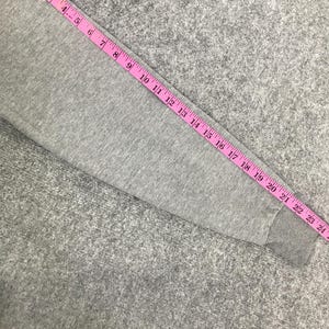 May include: A grey sweatshirt sleeve with a pink measuring tape laid across it. The tape shows measurements up to 61 cm. The sleeve is made of a soft, grey fabric.