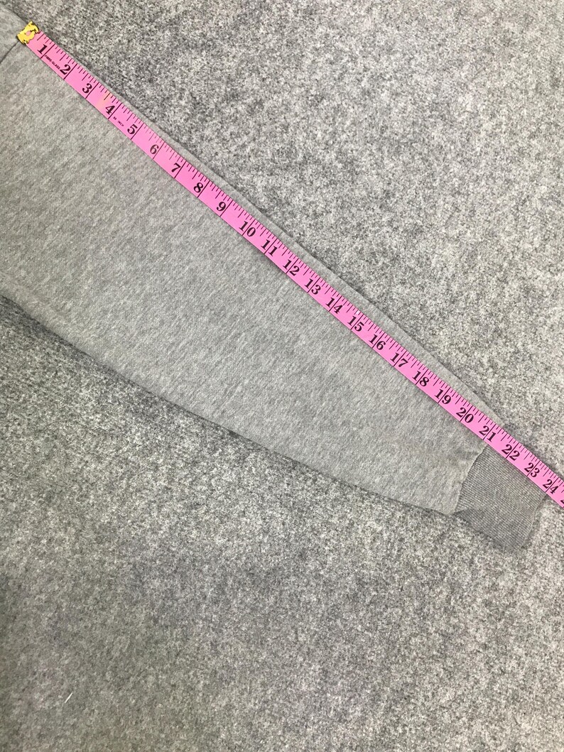May include: A grey sweatshirt sleeve with a pink measuring tape laid across it. The tape shows measurements up to 61 cm. The sleeve is made of a soft, grey fabric.