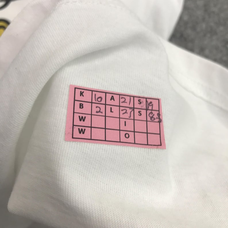 May include: A pink gridded tag with handwritten letters and numbers. The tag has the letters K, B, W, W and the numbers 10, 2, 2, 27, 19, 6.5. The tag also has the letters A, L, I, O and the numbers 2, 2, 1, 1.