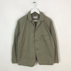 Puede incluir: Chaqueta verde oliva claro con cierre de botones, dos bolsillos delanteros y un pequeño bolsillo en el pecho. La chaqueta tiene cuello y mangas largas. Una etiqueta roja de Levi's es visible en el bolsillo del pecho.