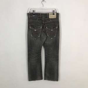May include: Faded black denim jeans with distressed details, featuring back pockets with red stitching and a leather patch. The jeans are hanging on a white hanger.