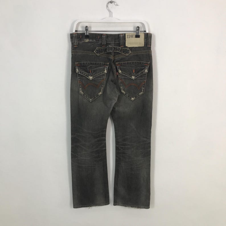 May include: Faded black denim jeans with distressed details, featuring back pockets with red stitching and a leather patch. The jeans are hanging on a white hanger.