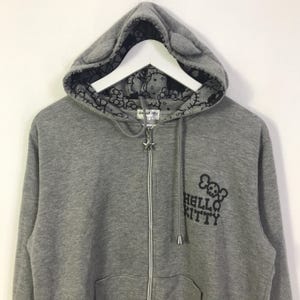 May include: Grey zip-up hoodie featuring a Hello Kitty skull graphic. The hood has cat ears and a black and white Hello Kitty pattern lining. The zipper pull is a star.