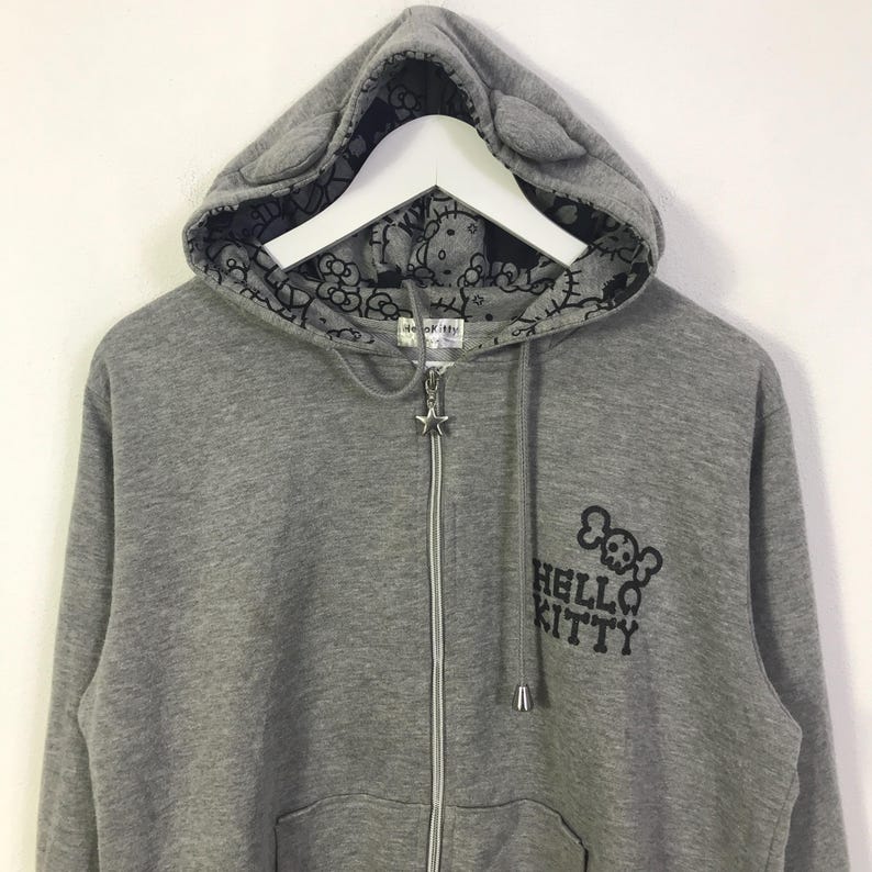 May include: Grey zip-up hoodie featuring a Hello Kitty skull graphic. The hood has cat ears and a black and white Hello Kitty pattern lining. The zipper pull is a star.