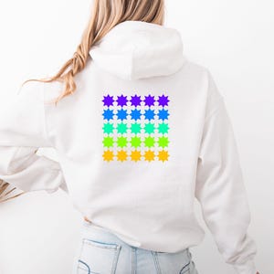 Könnte beinhalten: Weißer Kapuzenpullover mit einem quadratischen Muster aus farbenfrohen Sternen auf dem Rücken. Die Sternen platzen von Lila über Blau, Grün, Gelb bis Orange. Der Hoodie hat eine Kapuze und lange Ärmel.