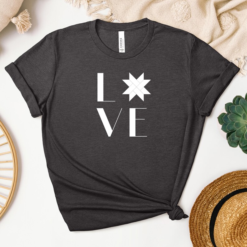 Puede incluir: Una camiseta de manga corta en color gris carb&oacute;n oscuro con el texto blanco LOVE impreso en la parte delantera. La letra O est&aacute; sustituida por un dise&ntilde;o geom&eacute;trico blanco de una estrella de ocho puntas. La camiseta tiene cuello redondo.