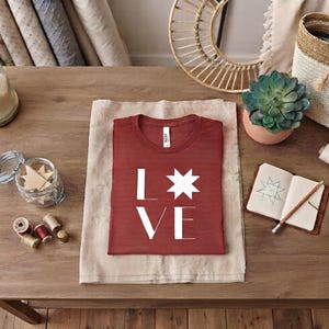 Puede incluir: Una camiseta de manga corta color &oacute;xido con el texto blanco LOVE, donde la letra O est&aacute; sustituida por un gr&aacute;fico de estrella blanca de ocho puntas. La prenda est&aacute; doblada cuidadosamente sobre una tela clara, mostrando un dise&ntilde;o sencillo.