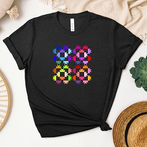 Könnte beinhalten: Schwarzes T-Shirt mit einem farbenfrohen, geometrischen Design. Das Design besteht aus vier Quadraten, die jeweils mit einem sternförmigen Muster in verschiedenen Farben gefüllt sind, darunter Blau, Lila, Rosa und Gelb. Das T-Shirt hat hochgekrempelte Ärmel.