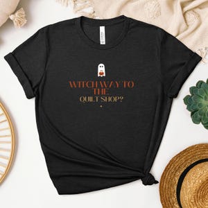 Könnte beinhalten: Schwarzes T-Shirt mit dem Text "Witch Way to the Quilt Shop?" in Orange und Weiß. Darüber befindet sich eine Geister- und Kürbisgrafik. Das T-Shirt ist am unteren Ende geknotet und gefaltet.