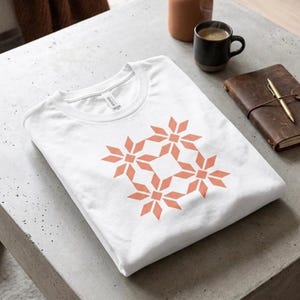 Könnte beinhalten: Ein weißes T-Shirt mit Rundhalsausschnitt und einem geometrischen Muster aus vier miteinander verbundenen korallenfarbenen Sternformen auf der Vorderseite. Das Shirt ist ordentlich gefaltet und zeigt die kurzen Ärmel sowie das Markenetikett.