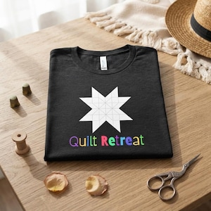 Könnte beinhalten: Ein schwarzes T-Shirt mit einem weißen geometrischen Sternmuster aus mehreren Dreiecken und dem mehrfarbigen Schriftzug Quilt Retreat unter dem Stern. Das T-Shirt hat einen Rundhalsausschnitt und ein sichtbares Markenetikett am Innenkragen.