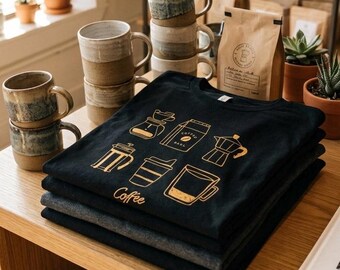 以下が含まれることがあります： 黒とダークグレーの折りたたまれたTシャツの山。一番上の黒いTシャツには、フレンチプレス、モカポット、コーヒードリッパーなど、さまざまなコーヒー抽出器具の金色のグラフィックプリントが施されており、その下にCoffeeという文字が印刷されています。