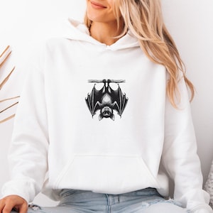 Könnte beinhalten: Weißer Kapuzenpullover mit einer schwarz-weißen Illustration einer Fledermaus, die kopfüber an einem Ast hängt. Der Kapuzenpullover hat eine Vordertasche und eine Kapuze mit Kordelzug. Die Flügel der Fledermaus sind detailliert.