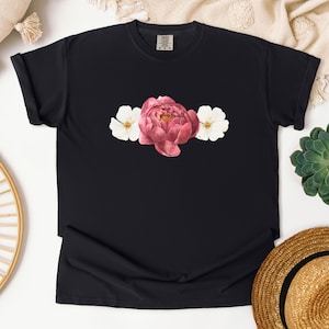 Könnte beinhalten: Schwarzes T-Shirt mit einem Aquarell-Blumenmuster. Das Design zeigt eine große rosa Pfingstrose in der Mitte, flankiert von zwei weißen Blumen. Das T-Shirt ist aus einem weichen, bequemen Material.