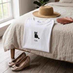 Könnte beinhalten: Weißes T-Shirt mit einer schwarzen Katzenzeichnung und dem Wort "Calm". Ein Strohhut und beige Keilabsatzschuhe sind ebenfalls zu sehen. Das T-Shirt liegt gefaltet auf einem Bett.